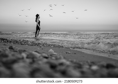 憂鬱と海。美しい少女は思慮深く海に見えます。創造的な不機嫌そうな芸術的なコンセプト。カモメが水の上を飛ぶ。黒のロングドレスと帽子。横向きの白黒コンポジション。一瞬