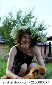 curly young girl in a wreath of wild flowers plays with a dog in the yard of a country house