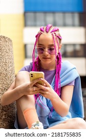 Retrato de uma adolescente caucasiana hipster com tranças rosas usando um smartphone em um fundo de rua. Conceito de verão. Estilo Geração Z. Conceito de mídia social. Foco seletivo.