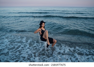Beautiful brunette woman in black dress and hat sitting on white chair in sea waves water. Horizontal composition sunset time soft blue and pink golden hour light. Creative artistic summer moment