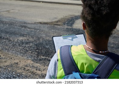 wanita muda kulit hitam memeriksa pekerjaan aspal yang baru diaplikasikan dengan mesin berat di jalan yang telah direnovasi sambil membuat laporan untuk perusahaan