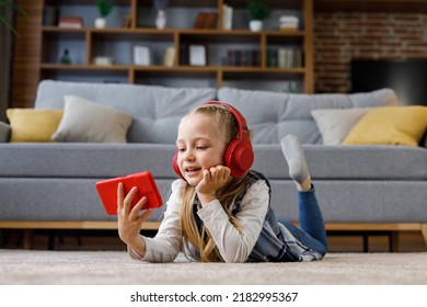 Gadis kecil mengenakan earphone merah tergeletak di karpet di rumah. Anak lucu tersenyum memegang smartphone, menonton kartun, browsing internet atau menonton video di media sosial, bermain game online
