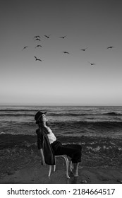 girl in a formal elegant stylish suit, shirt and hat sitting on a chair in the waves of the sea. artistic dreamer portrait of Creative black and white composition. birds in the sky. calm before storm