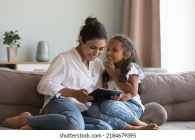 Happy Indian little daughter kid talking to mom, using digital tablet, sitting on sofa at home, smiling, laughing. Mother and child enjoying leisure, playing game on digital gadget