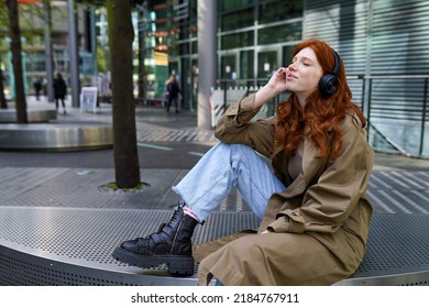 Happy teen stylish redhead hipster girl model sitting on urban street wearing headphones listening to music with eyes closed feeling relaxed and serene in big city rush outdoors.