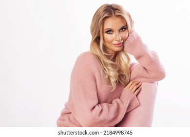 ピンクのセーターを着た若い美しいブロンドの髪の女性が、ピンクの肘掛け椅子に座り、笑顔でカメラを見ています。女性の肖像画。白で隔離されます。