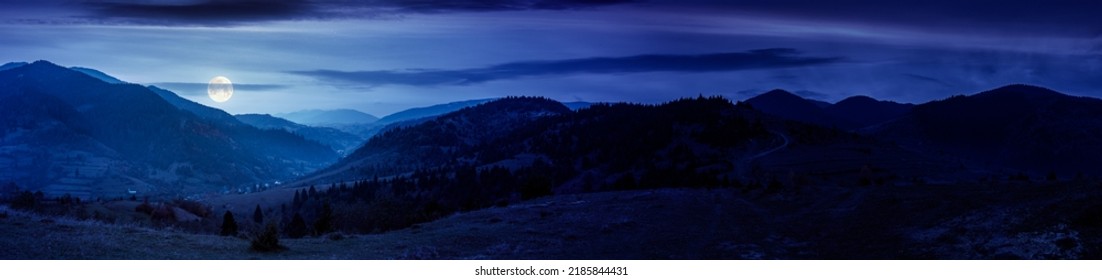 panorama del paisaje rural por la noche. prados de pastos herbosos y colinas boscosas en otoño. cresta de la montaña en la distancia. pueblo en el valle. nubes en el cielo a la luz de la luna llena