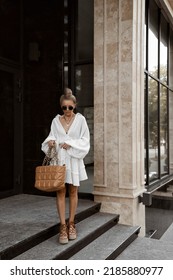 beautiful blonde girl dressed in white oversized magnificent dress, brown lace-up sandals, bag, accessories, sunglasses, bundle hairstyle, stylish summer fashion outlook, full length lifestyle model