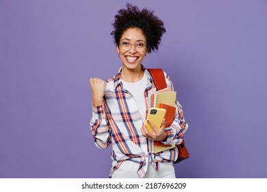 Junge lustige Mädchen Frau der afroamerikanischen Ethnizität Teenager-Studentin im Hemdrucksack benutzt das Handy und macht eine Gewinnergeste, die auf einem schlichten lila Hintergrund isoliert ist. Bildung im High School College-Konzept