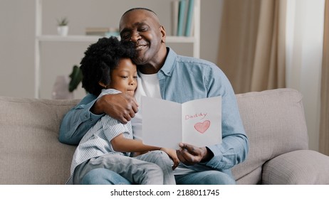 Reife afroamerikanesche Mann mat léiwem Kand Meedchen, dat am Zëmmer um Sofa sëtzt, Papp liest Gréisskaart, léif Duechter huet e Kaddo mat eegenen Hänn fir Gebuertsdag oder Pappendag gemaach, Papp glécklech gekuschelt