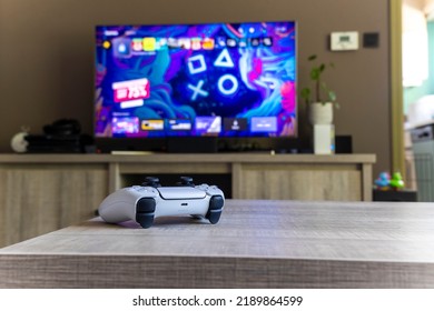 BRECHT, BÈLGICA – 14 D'AGOST DE 2022: un retrat d'un controlador sense fil bàsic blanc de Sony Playstation 5 sobre una taula de fusta davant d'un televisor que mostra la pantalla de la Playstation Plus PS5 en una sala d'estar