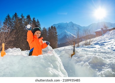Cậu bé hạnh phúc ném quả cầu tuyết đứng trong pháo đài tuyết với nền núi Alps trong kỳ nghỉ mùa đông