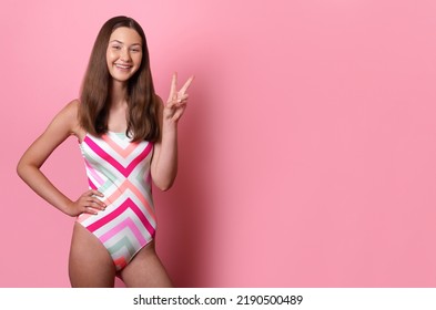 Une adolescente souriante et joyeuse en maillot de bain une pièce à rayures colorées et lumineuses montre un signe de paix isolé sur fond rose, une bannière avec un espace libre à droite