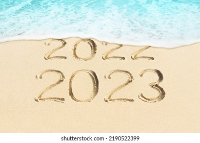 Foto konsep Tahun Baru. Teks 2023 dan 2022 tulisan tangan di permukaan pasir. Gelombang laut biru menghanyutkan angka-angka di pantai.