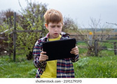 Anak laki-laki berambut merah yang bahagia bermain tablet atau menonton kartun, berjalan di halaman belakang desa, liburan musim panas, gaya hidup.