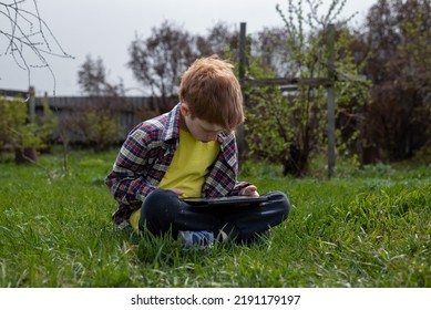 Anak laki-laki berambut merah yang bahagia bermain tablet atau menonton kartun sambil duduk di rumput hijau di halaman belakang desa, liburan musim panas.