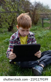 Anak laki-laki berambut merah yang bahagia bermain tablet atau menonton kartun sambil duduk di rumput hijau di halaman belakang desa, liburan musim panas, close-up.