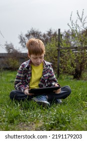Anak laki-laki berambut merah yang bahagia bermain tablet atau menonton kartun sambil duduk di rumput hijau di halaman belakang desa, musim panas.