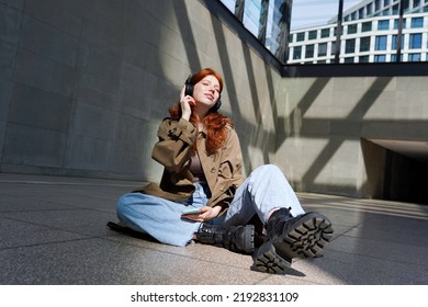 Happy teen redhead girl hipster model sitting near urban walls background wearing wireless headphones listening to mobile music with eyes closed enjoying streaming service app outdoors.