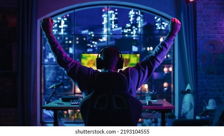 Foto de la parte posterior de la cabeza de un jugador afroamericano que gana un videojuego en una computadora personal en una sala de estar iluminada con luces de neón en su casa. Una tarde acogedora en un apartamento tipo loft.