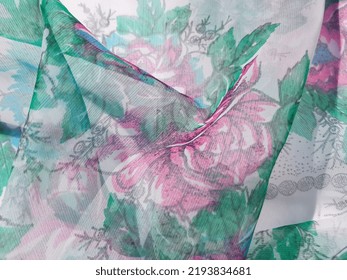 Tejido ligero ondulado translúcido ligeramente brillante (organza) con estampado floral de siluetas de flores rosas y azules en hojas verdes; con borde gris, en pliegues (macro, textura).