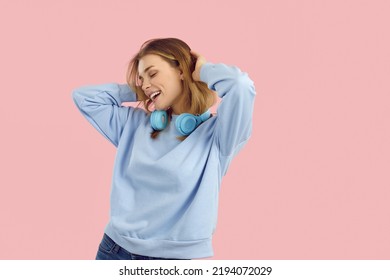 Portrait of young woman wearing headphones around her neck, dancing and having fun on pink background. Beautiful charming smiling girl in light blue casual sweater and jeans. People lifestyle