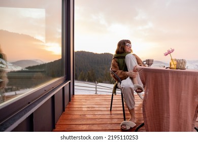 Mujer sentada en la terraza con una gran vista de las montañas en invierno, disfrutando de una bebida y un hermoso paisaje durante el amanecer. Concepto de descanso y escape a la naturaleza.