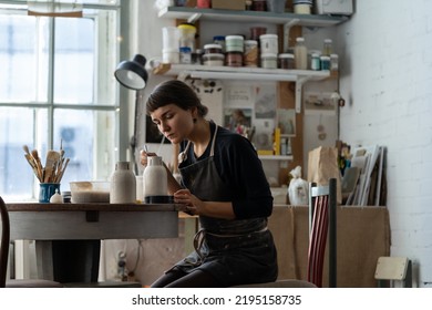 Young female artisan in black apron works with crockery sitting at round table in workshop against bright window. Brunette woman enjoys painting handmade ceramic vase in pottery craft studio