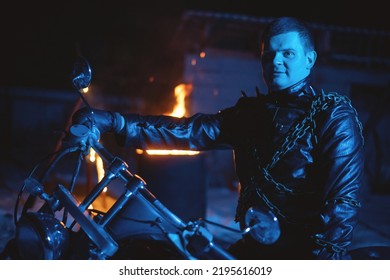 Motorbiker in the black leather jacket with the spikes in the blue light on the burning fire background.