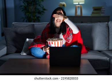 Femeie hispanică care mănâncă floricele de porumb, vizionează un film pe canapea, zâmbind și râzând cu mâna pe față acoperind ochii pentru surpriză. concept orb.