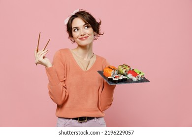 Mladá zamyšlená zamyšlená zábavná žena ve svetru drží v ruce makizushi sushi rolka podávaná na černém talíři tradiční japonské jídlo odhlédneme od hůlek izolovaných na obyčejném pastelově růžovém pozadí.