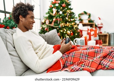 Young african american man playing video game sitting on the sofa by christmas tree at home.