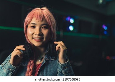 Teenage gamer girl with pink short hair wearing headset on her neck looking into camera smiling with gaming setup in the background. High quality photo