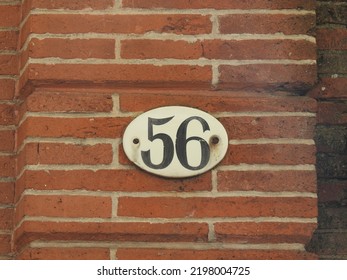 House number (address) fifty six (56) sign, black numbered white plate (closeup) against red (orange) bricks wall of old building background. Grunge, texture, wallpaper