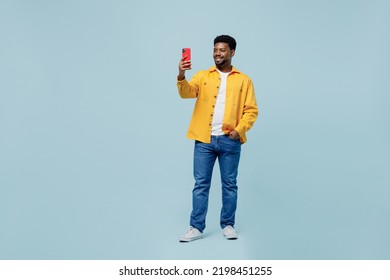 Full body young man of African American ethnicity wear yellow shirt doing selfie shot on mobile cell phone post photo on social network isolated on plain pastel light blue background studio portrait.
