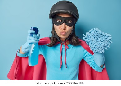 Verwonderde vrouw gekleed in superheldenkostuum houdt lippen gevouwen houdt schoonmaakmiddel en doek vast doet huishoudelijke klusjes draagt ​​kostuum met mantel geïsoleerd over blauwe achtergrond. Huishoudelijk werkconcept