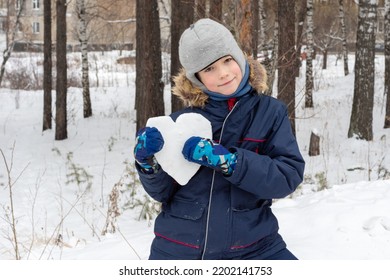 Kinderen winter. Schattige lachende jongen in een winterjas en wanten met een besneeuwde hartvorm op straat na een sneeuwval. Sneeuwhart in de handen. Valentijnsdag, liefdesconcept.