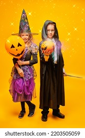 ハロウィンマジック。魔女と魔法使いの衣装を着た 2 人の幸せな子供が、ハロウィーンの風船とカボチャのバスケットを持っています。周りに魔法の光がある黄色の背景。トリック・オア・トリート。