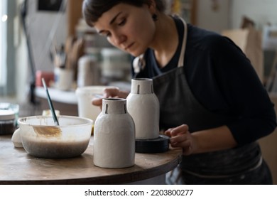 Una joven artesana con delantal negro trabaja con vajilla sentada en una mesa redonda en un taller contra una ventana luminosa. La mujer morena disfruta pintando un jarrón de cerámica hecho a mano en un estudio de artesanía de cerámica