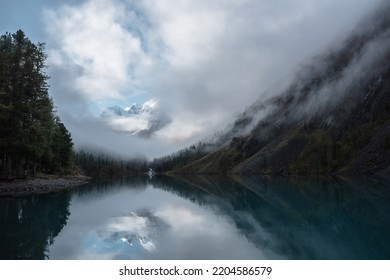 雲の中に雪の城がある静かな風景。マウンテン クリークは、森の丘から氷河湖に流れ込みます。霧が立ち込める雪山、静かな高山湖に映る小さな川と針葉樹。