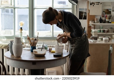 Young female artisan stands near round wooden table with equipment for work with pottery against bright big window. Brunette woman in black apron pours white paint into ceramic cup in workshop