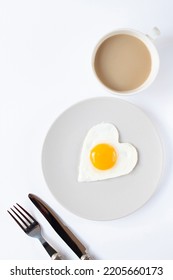 Un huevo frito en forma de corazón en un plato, una taza de café y cubiertos están sobre un fondo claro. Día de San Valentín. Una imagen vertical.