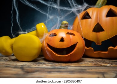 Yellow gym dumbbells and ceramic Halloween Jack O Lantern figurine pumpkins, covered with spider web. Healthy fitness lifestyle autumn or fall composition. Gym workout and sport training concept.