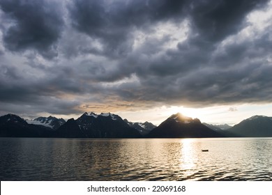 Sonnenuntergangslandschaft mit dunklen Wolken über einem See mit schneebedeckter Bergküste