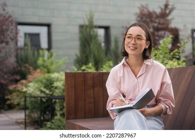 Portrait of confident smiling asian writer taking notes looking away sitting in park. Smart student wearing eyeglasses studying, reading book sitting in university campus. Education concept