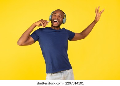Cantante di talento di colore che canta una canzone tenendo in mano uno smartphone come un microfono, indossando auricolari wireless, in posa con gli occhi chiusi, in piedi su uno sfondo giallo da studio. Concetto di applicazione musicale