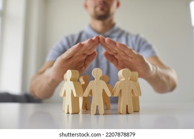 Junger Mann hält Hände über kleinen hölzernen Spielzeugfiguren auf weißem Schreibtisch als Metapher für den Schutz der Menschenrechte und eine sichere Gemeinschaft von Menschen. Nahaufnahme, Nahaufnahme. Gesellschaft, Pflege, Sicherheitskonzept