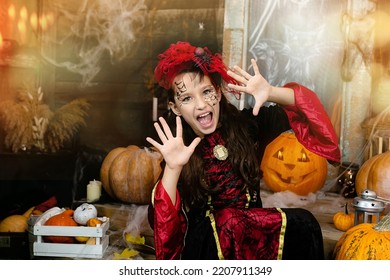 Halloween holiday concept. Portrait of child in wearing black witch costume, makeup, scares in carnival night on wooden background, surround by web, carved pumpkins. Girl having fun, trick or treat.