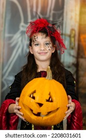 Portrait smiling child in witch costume, headwear, face painted halloween makeup. Caucasian teenager girl holding orange carved pumkin. Kids trick or treating. Concept of holiday, Seasonal carnaval