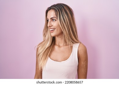 Junge blonde Frau steht vor rosa Hintergrund und schaut zur Seite, mit einem Lächeln im Gesicht und natürlichem Ausdruck. Lachend selbstbewusst.